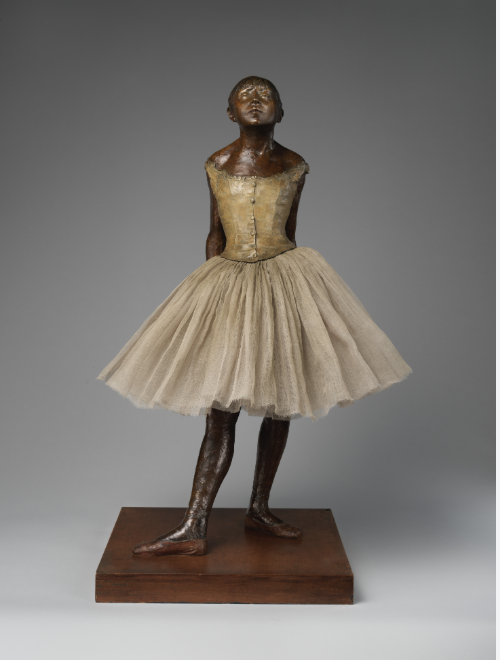 A bronze ballerina with a light tutu stands on a wooden base at the Met, arms behind her back, head tilted upward.